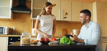 Attractive couple chatting in the kitchen early morning. Handsome man using tablet while his girlfriend cooking breakfast at home