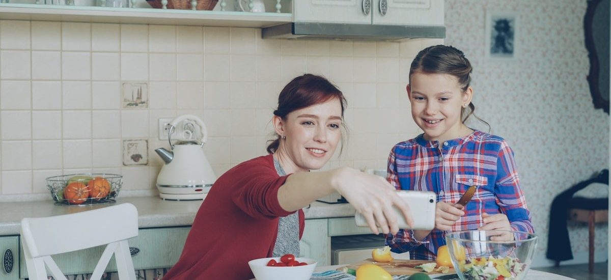 Smiling mother making selfie portrait together with young cute daughter cooking breakfast at home in kitchen. Family, cook, and people concept