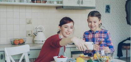 Smiling mother making selfie portrait together with young cute daughter cooking breakfast at home in kitchen. Family, cook, and people concept