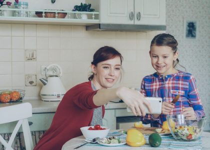 Smiling mother making selfie portrait together with young cute daughter cooking breakfast at home in kitchen. Family, cook, and people concept